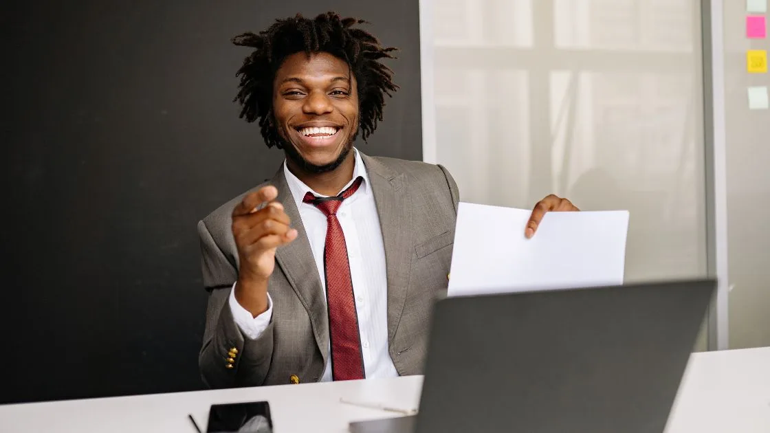 A smiling small business owner at a desk with a laptop, holding a printed marketing plan