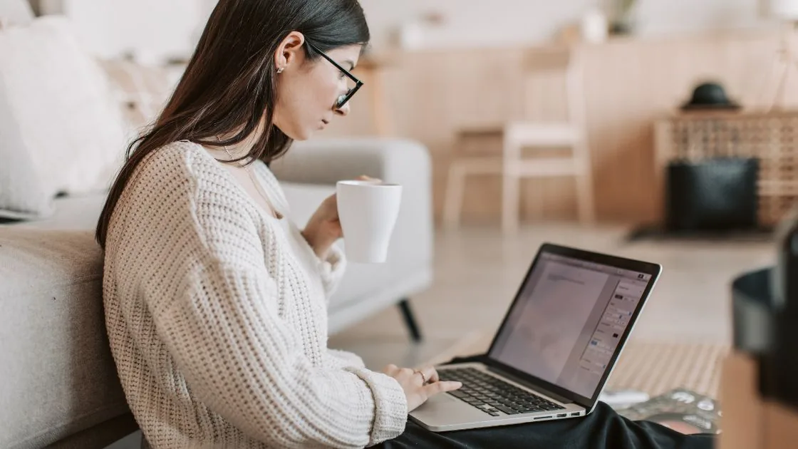 A small business owner in a knit sweater holding a coffee mug while working on a laptop at home