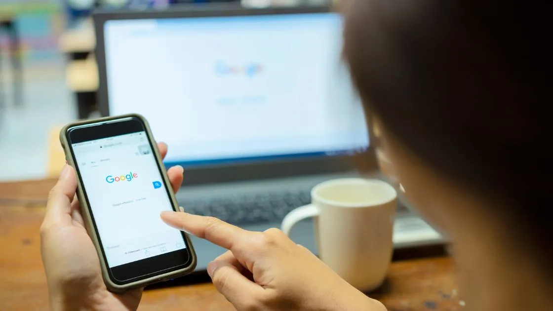 Person searching Google on a smartphone in front of a laptop showing the Google homepage